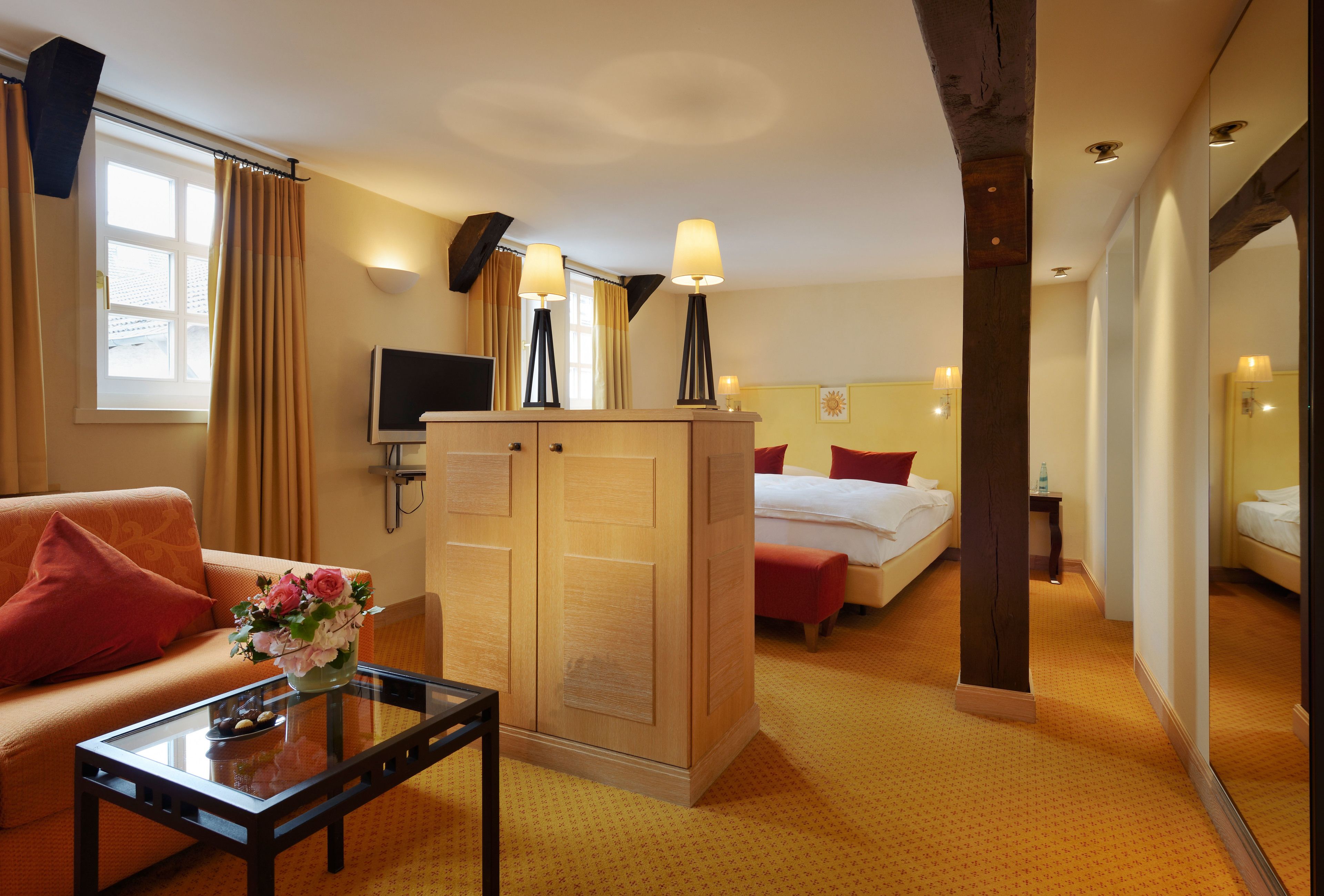 A hotel room with a sofa, a glass coffee table with a vase, a wooden wardrobe, a bed with red pillows, and a bedside table with a lamp.