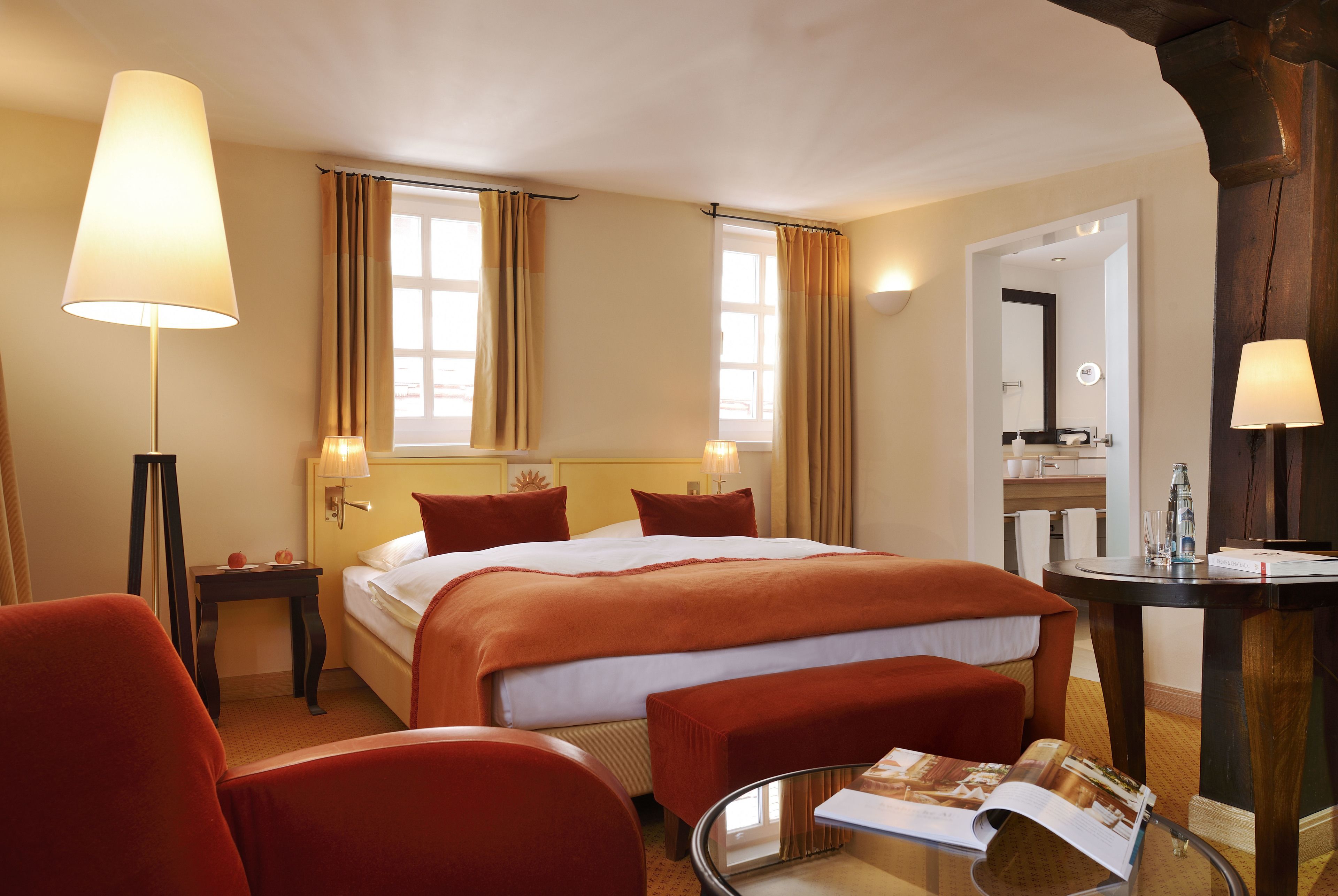 Een hotelkamer met een groot bed, twee bedlampen, een ronde tafel met een spiegel en een rode fauteuil.