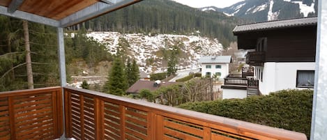 A wooden balcony with a view of a mountainous landscape and a white building with a dark roof.