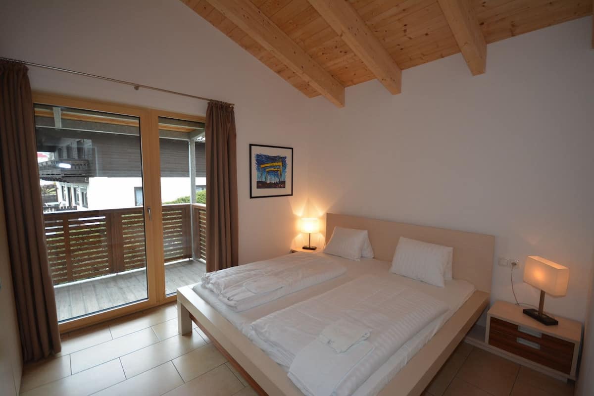 Une chambre avec un grand lit, deux tables de chevet avec des lampes, une porte-fenêtre coulissante donnant sur un espace extérieur et un tableau encadré au mur.