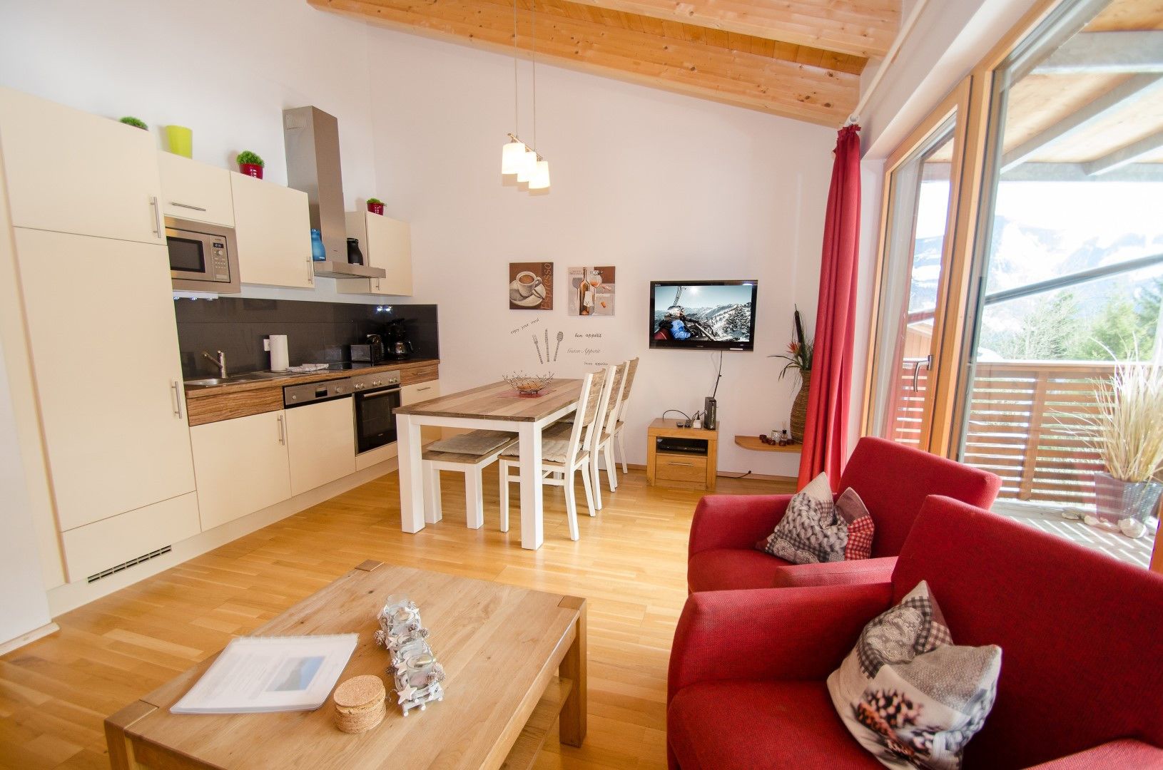 A modern living room with a red sofa, wooden coffee table, and a dining area with a wooden table and chairs. A kitchen with white cabinets and a microwave is visible in the background.
