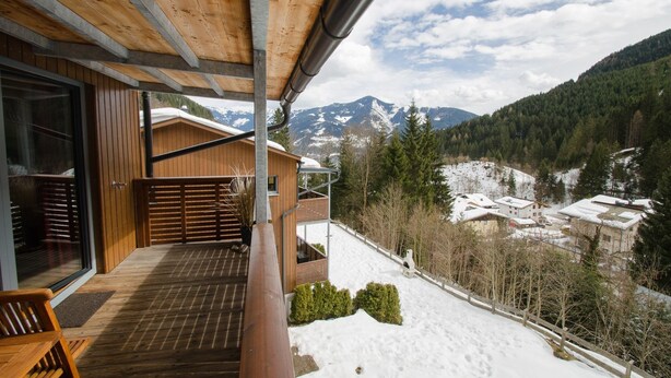 A wooden deck with a view of snow-covered mountains and a village. - Schmitten Finest Apartments (Zell am See)