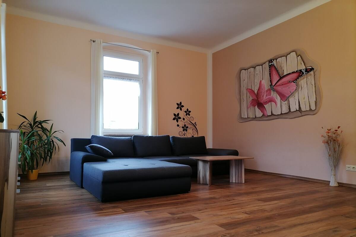 A living room with a sectional sofa, a small wooden table, a potted plant, and a wall art piece.