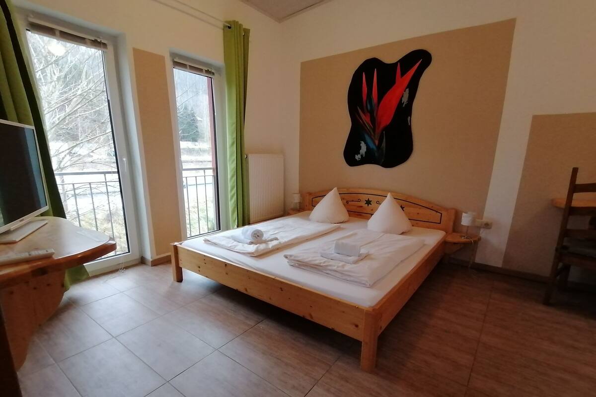 A neatly made bed with pillows and a rolled towel, a wooden desk with a computer, a chair, and a wall-mounted artwork.