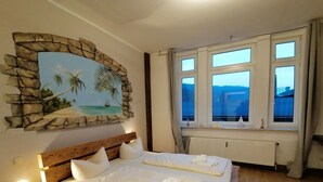 A bedroom with a wooden bed, a stone-framed painting of a beach scene, and a window with a view of a building. - Pension Bomätscher (Koenigstein)