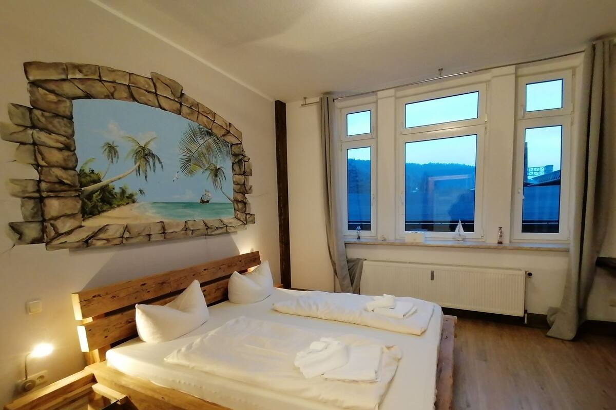 A bedroom with a wooden bed, a stone-framed painting of a beach scene, and a window with a view of a building.