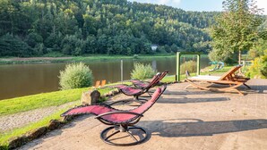 A lakeside patio with lounge chairs and a view of a forested hillside. - Pension Bomätscher (Koenigstein)