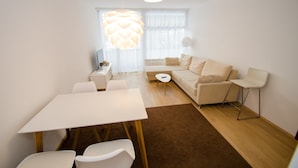 Een moderne woonkamer met een bank, een eettafel met stoelen en een groot raam met dunne gordijnen.