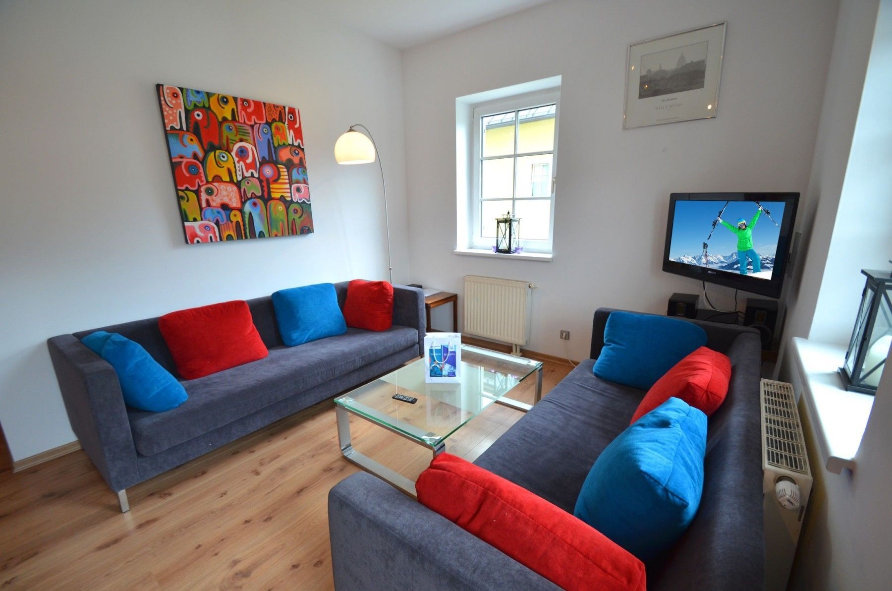 A modern living room with two grey sofas, colorful cushions, a glass coffee table, a TV, a window with a view, and a painting on the wall.