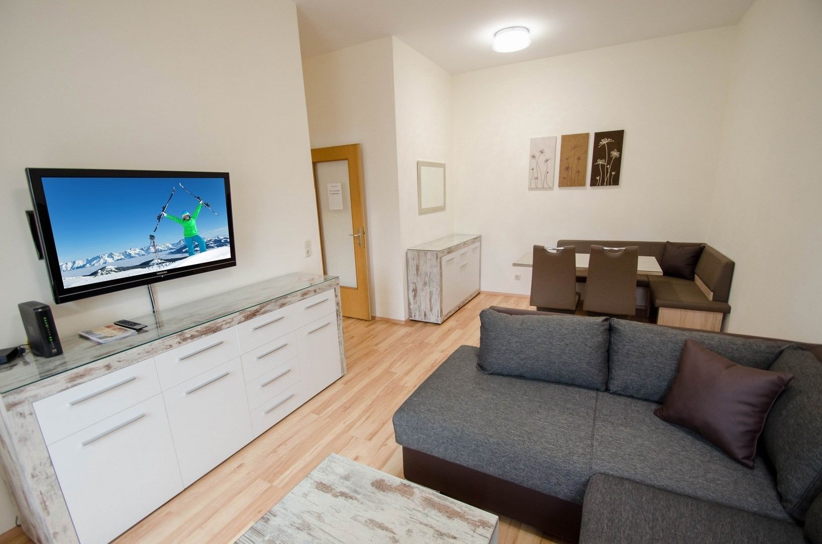 Uma sala de estar moderna com uma televisão de tela plana, um sofá cinza, uma mesa de centro de madeira e um armário branco.