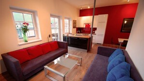 A modern living room with a brown sofa, a glass coffee table, and a purple sofa with blue cushions. A kitchen with red walls and white cabinets is visible in the background. - Apartments CityXpress (Zell am See)