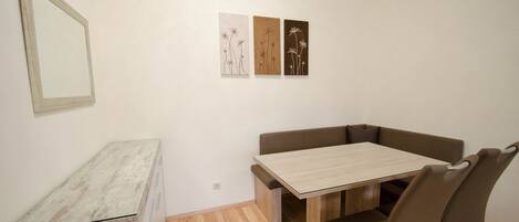 Een eethoek met een houten tafel, beklede bruine stoelen, een klein wit kastje en drie kunstwerken aan de muur.