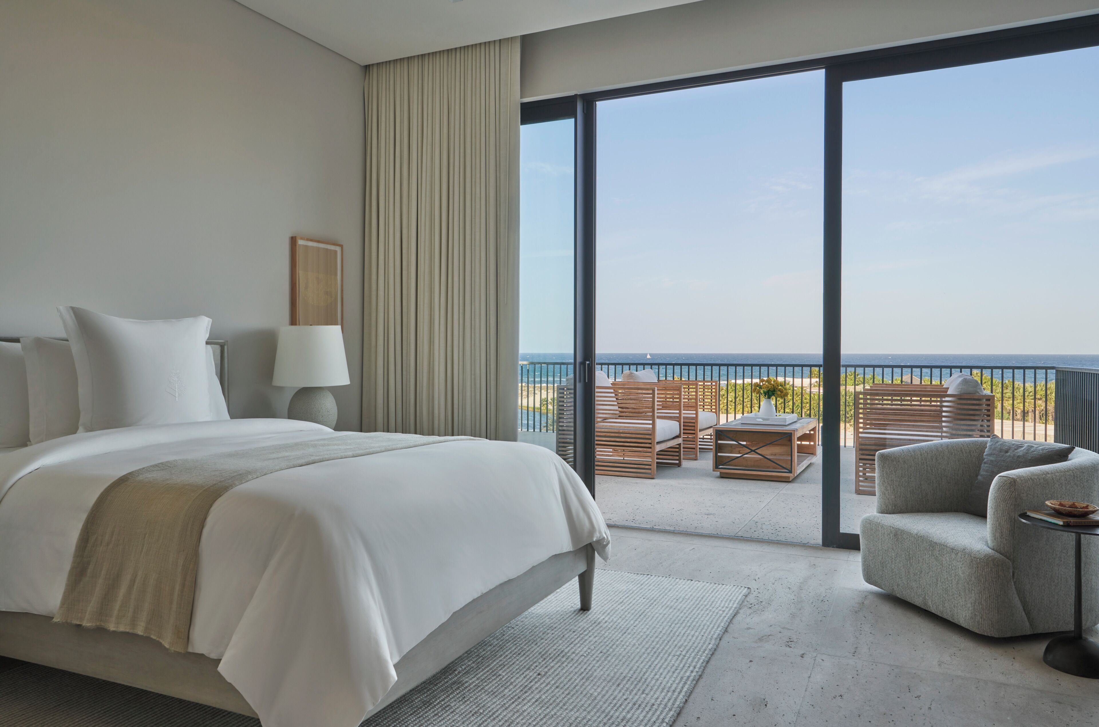 A hotel room with a large bed, a sitting area with a view of the sea, and a balcony with outdoor furniture.