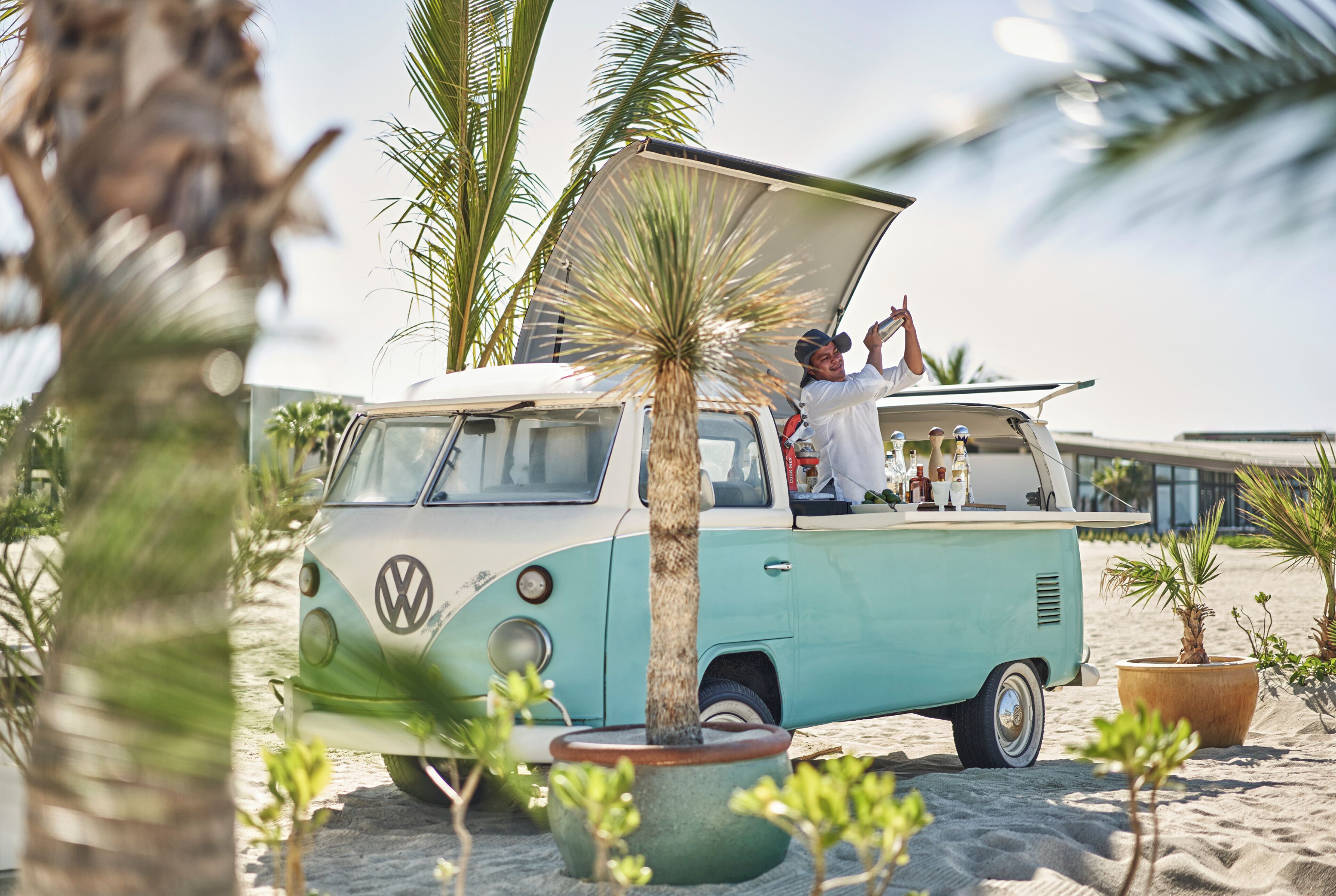 Un camper Volkswagen vintage parcheggiato su una spiaggia sabbiosa, con una palma sullo sfondo.