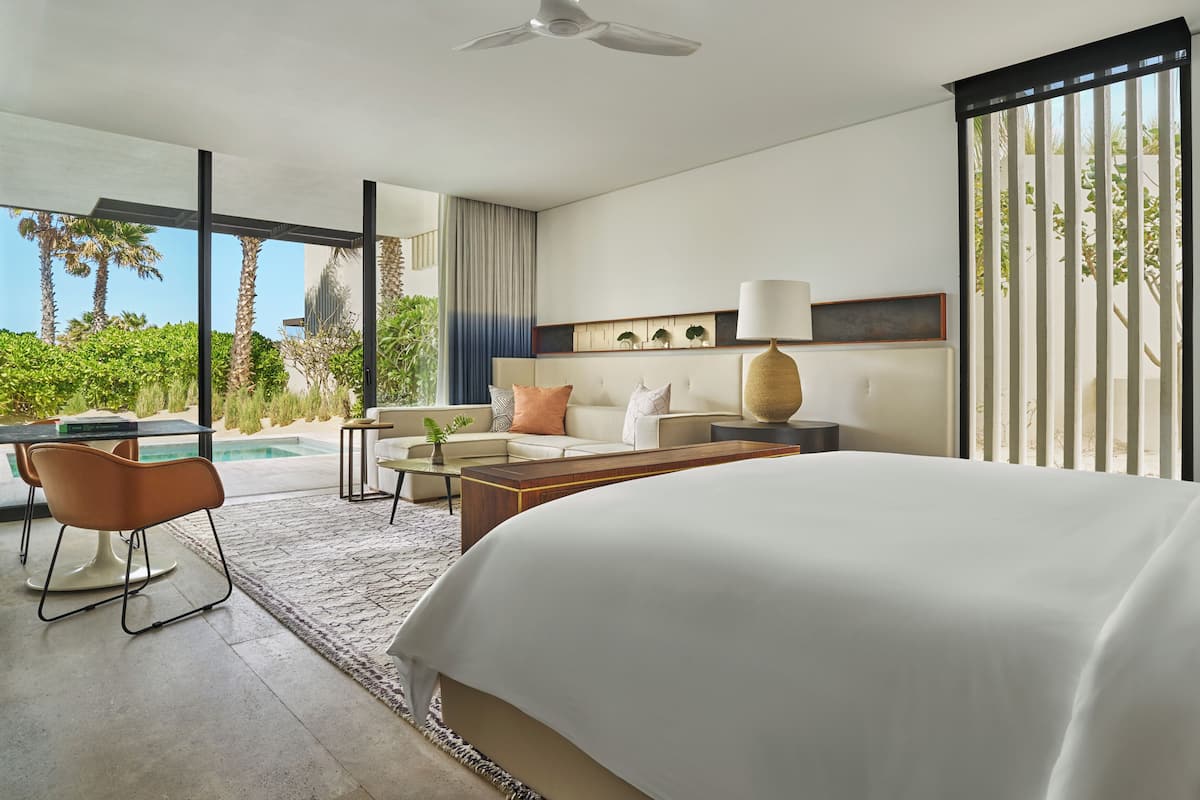 A modern hotel room with a large bed, a desk with a chair, a sofa, and a view of a pool and palm trees.