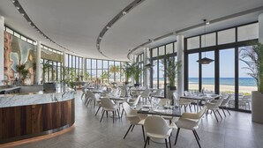 A modern restaurant with a curved counter, white chairs, and large windows offering a view of the beach.