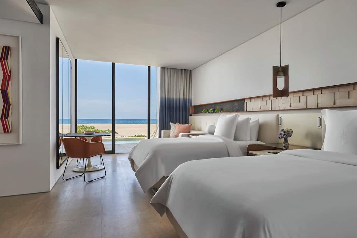 A modern hotel room with two beds, a dining area with a view of the beach, and a large window.