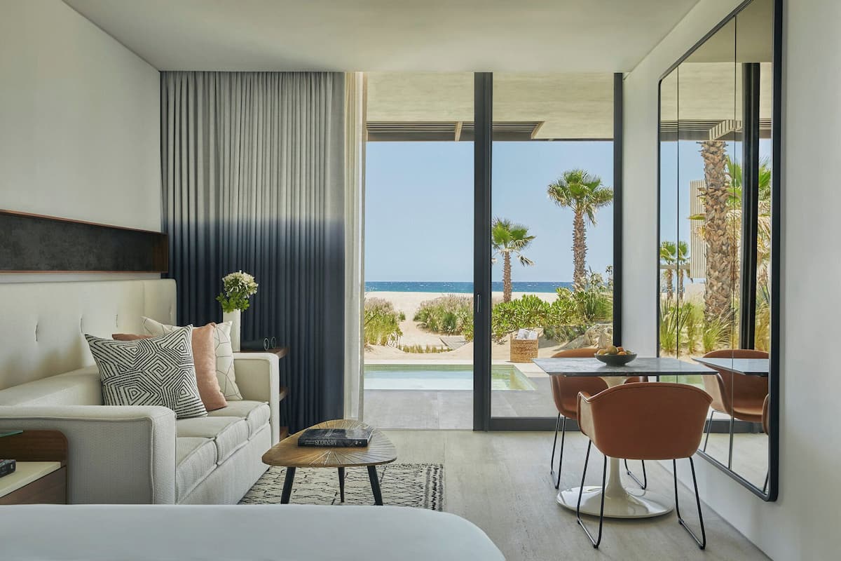 A modern hotel room with a large balcony, a sofa, a dining table, and a view of the beach and ocean.