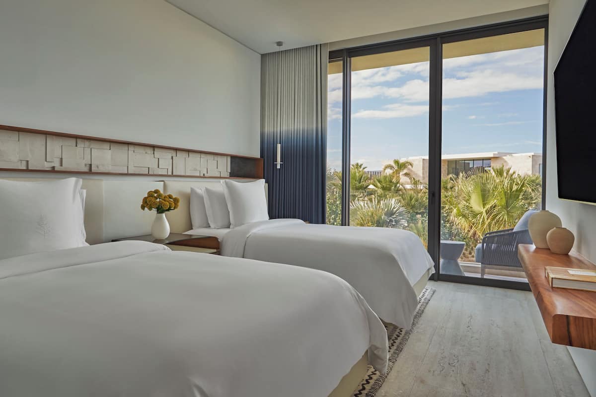 A hotel room with two beds, a nightstand, a vase with flowers, and a view of palm trees and buildings.
