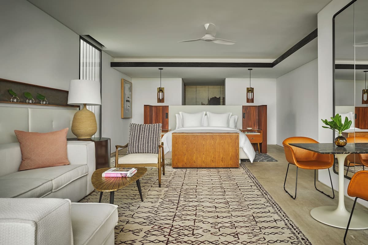 A modern hotel room with a large bed, a sofa, a dining table with chairs, a coffee table, and a large mirror.