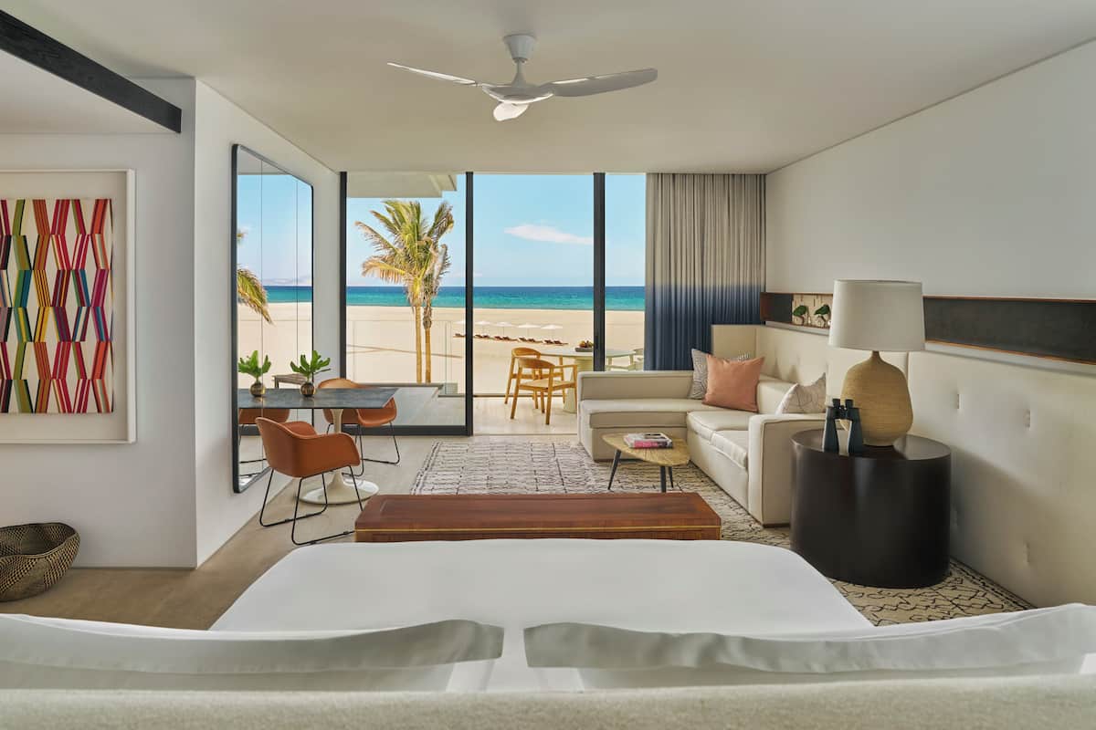 A hotel room with a bed, a seating area with a table and chairs, a sofa, a lamp, and a view of the beach.