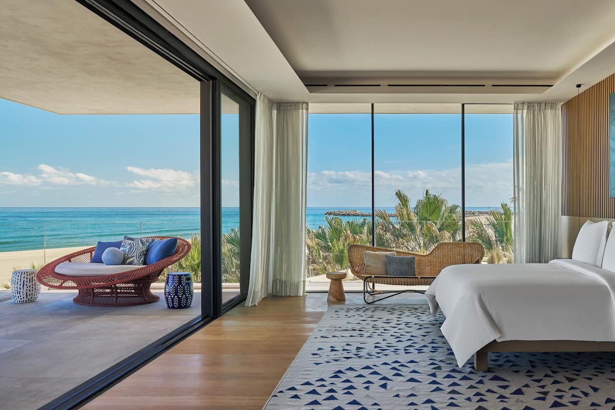 A modern hotel room with a large bed, a seating area with a wicker chair, and a view of the ocean.