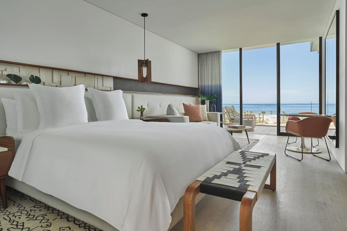 A modern hotel room with a large bed, a bedside table, a nightstand, a bench, a chair, and a view of the ocean.