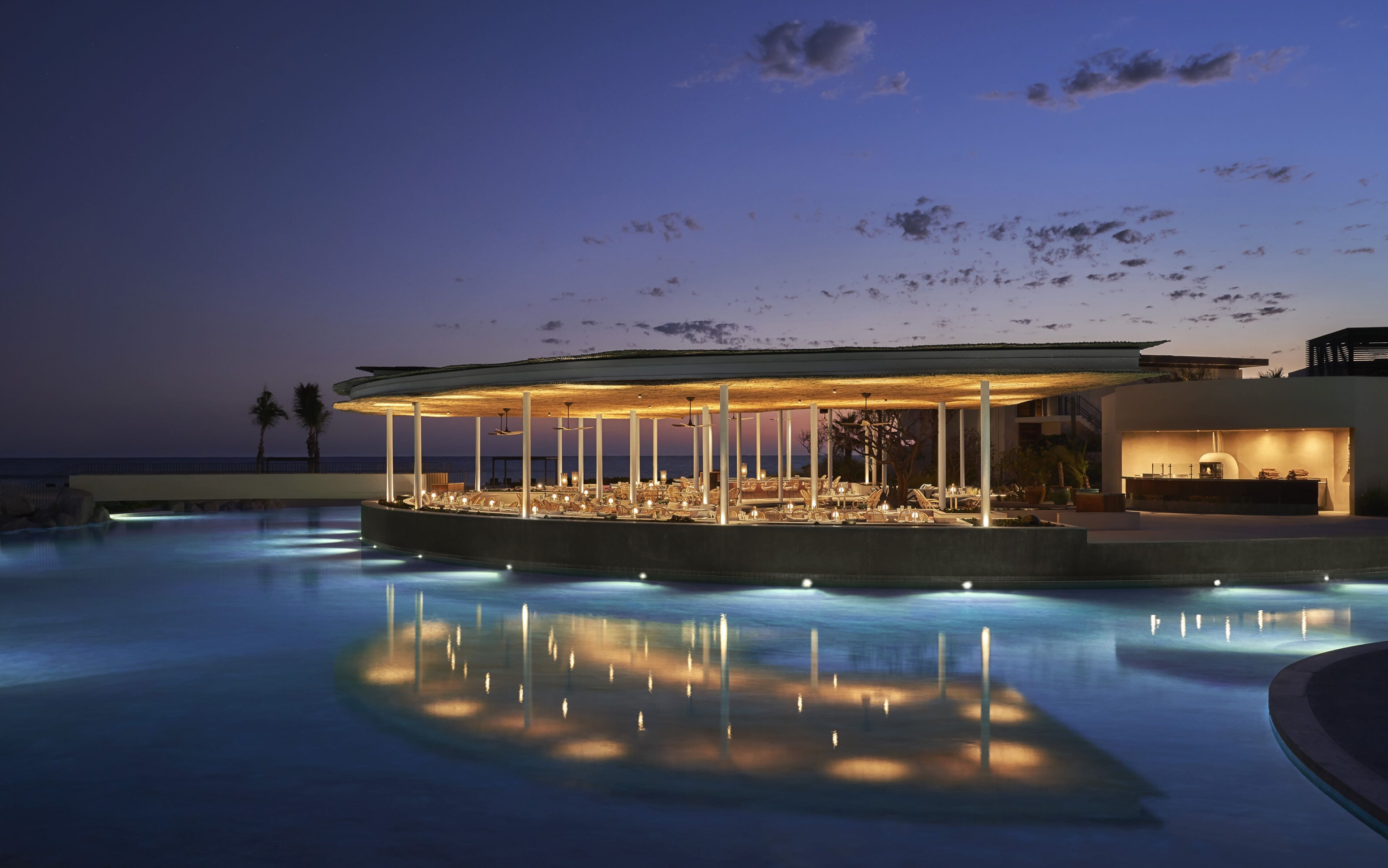 Zona de descanso junto a la piscina con un área de estar cubierta, una piscina que refleja luces y vistas al océano.
