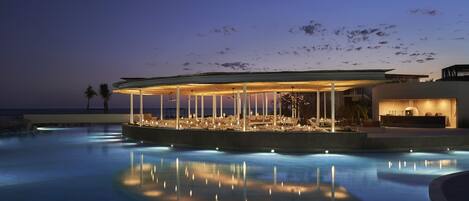 Zona de descanso junto a la piscina con un área de estar cubierta, una piscina que refleja luces y vistas al océano.