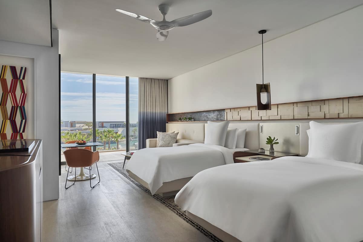 A hotel room with two beds, a desk, a chair, a ceiling fan, and a large window with a city view.