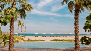 A beach resort with a pool, palm trees, beach chairs, and umbrellas.