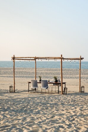 A beach setting with a wooden pergola, two chairs, a table with a potted plant, and string lights. - Four Seasons Resort Los Cabos at Costa Palmas (La Ribera)