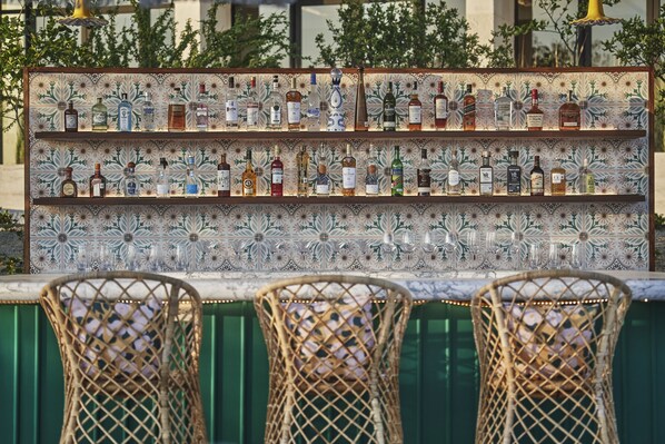 Un espace bar avec un mur recouvert de carreaux où sont exposées différentes bouteilles d’alcool, des chaises en osier et un comptoir vert.