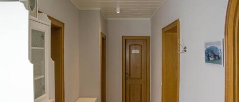 Un pasillo estrecho con una puerta de madera al final, un armario blanco con cajones y una pequeña imagen en la pared.