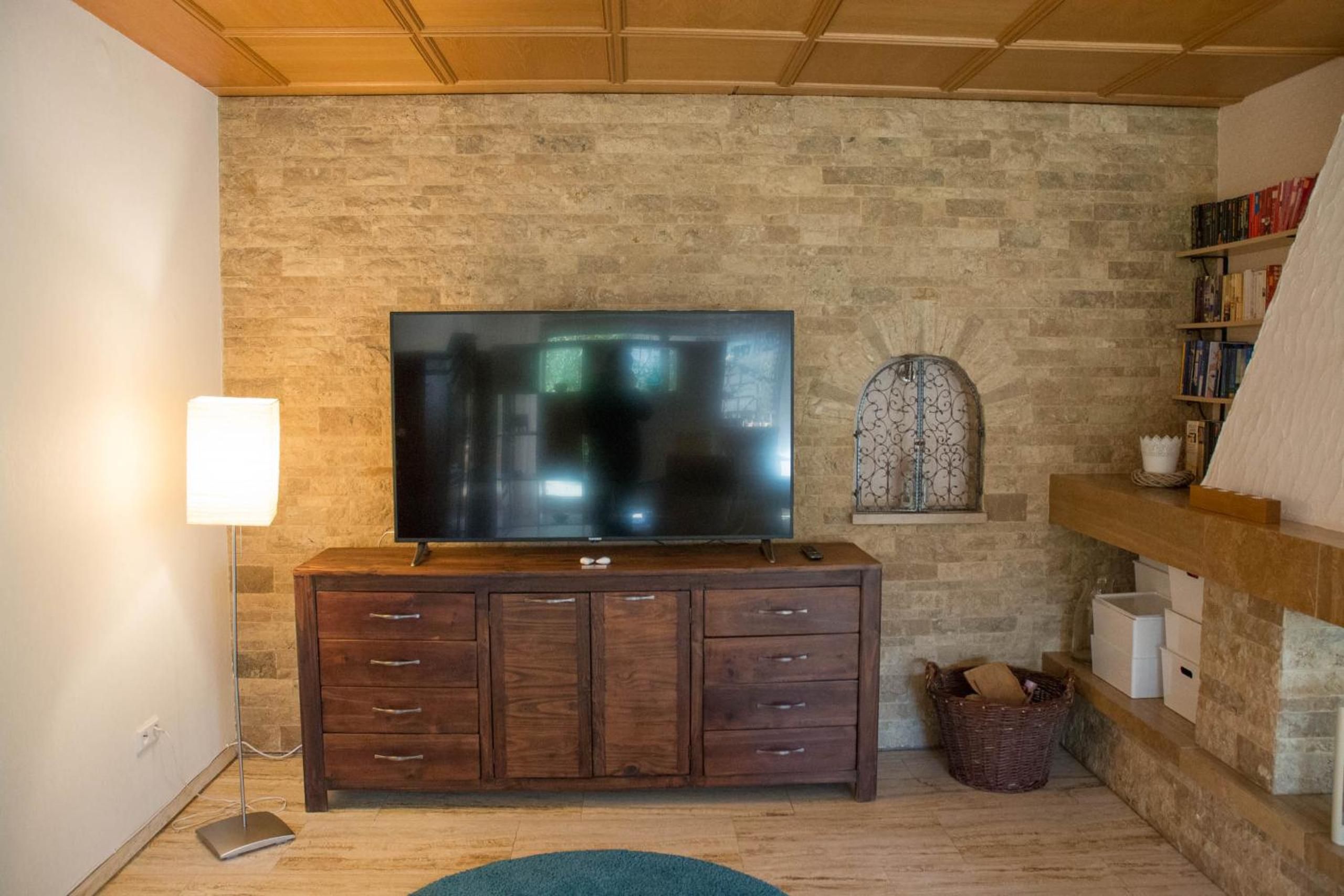 A living room with a stone wall, a flat-screen TV on a wooden cabinet, a floor lamp, a small decorative window, and a bookshelf.