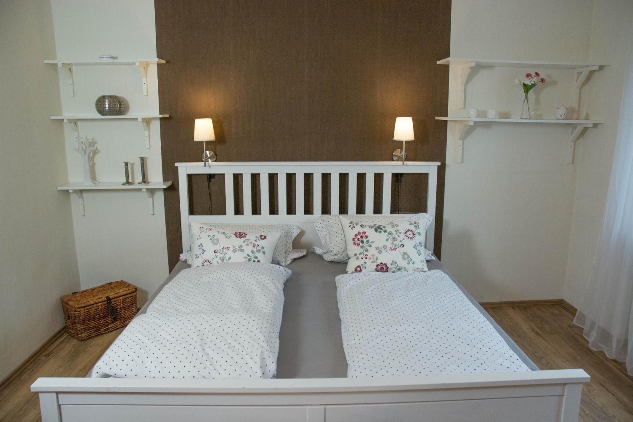 A double bed with a white headboard and two bedside lamps, flanked by white shelving units with decorative items.