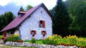 Une maison en pierre avec un toit en pente, deux fenêtres avec volets et un jardin où poussent des tournesols.