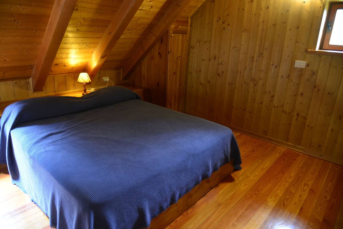 Een kamer met een houten plafond, een bed met een blauwe spreidekking, een nachtkastje met een lamp en een raam met uitzicht op het buiten.
