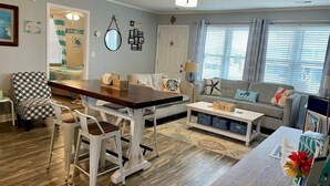 Ein Wohnzimmer mit einem Holzesstisch, zwei Barhocker, einer Couch und einem Couchtisch. Eine Tür führt in einen anderen Raum, und ein Fenster mit Vorhängen ist vorhanden.