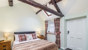 A bedroom with a sloped ceiling, a bed with a patterned blanket, two bedside lamps, a nightstand, a window with plaid curtains, and a white door. - Stilworth House (York)