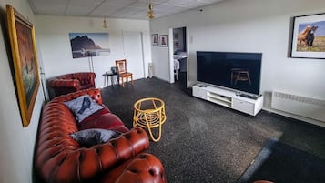 A living room with a large flat-screen TV, a red leather sofa, a wicker chair, and a small table.