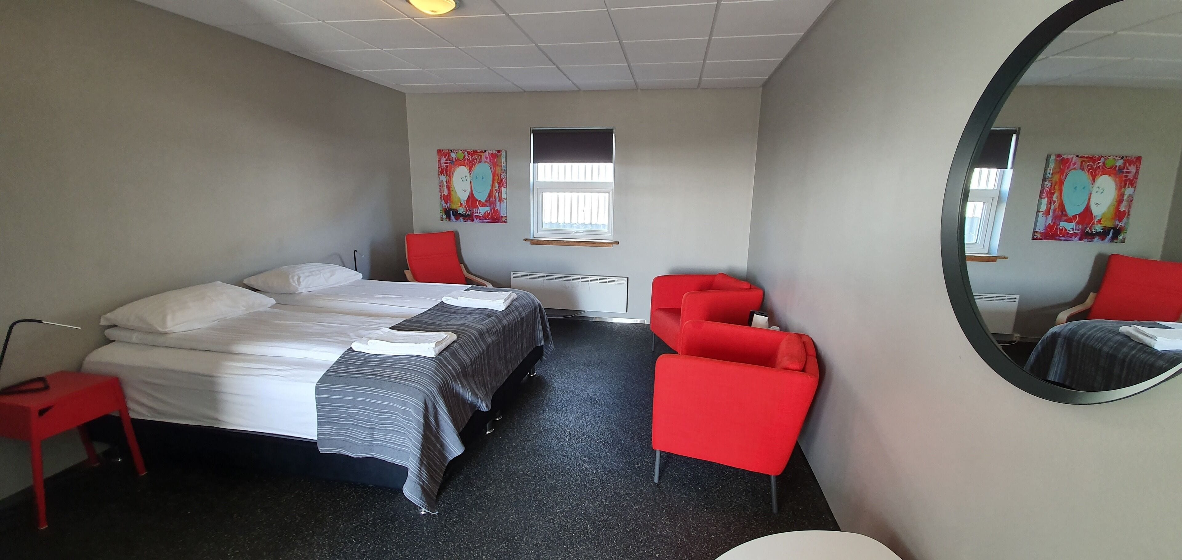 Una habitación de hotel con una cama, dos sillas rojas, un espejo redondo y obras de arte en las paredes.