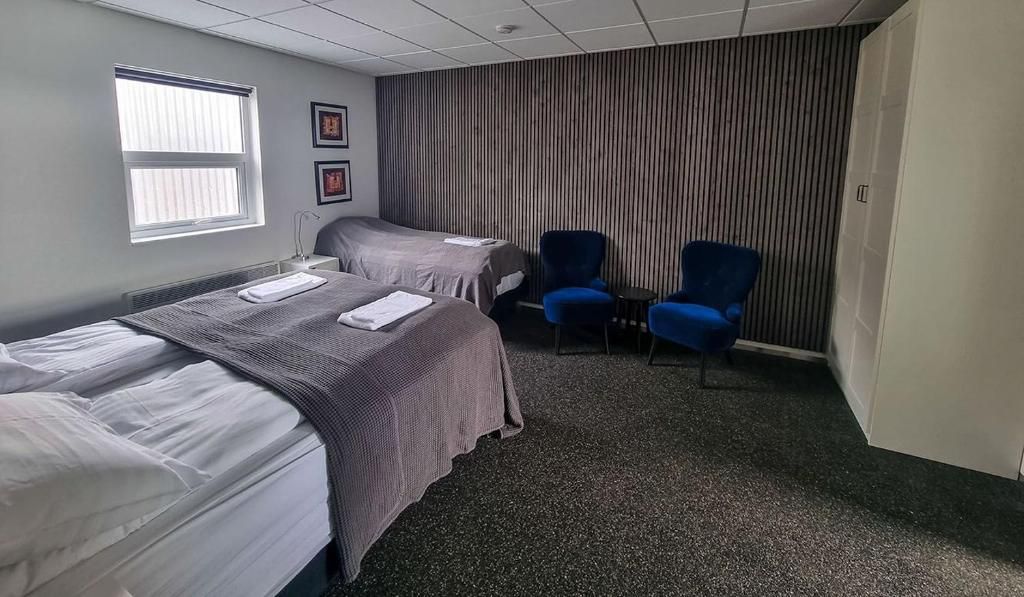 A hotel room with two beds, a window, a radiator, and two blue chairs.