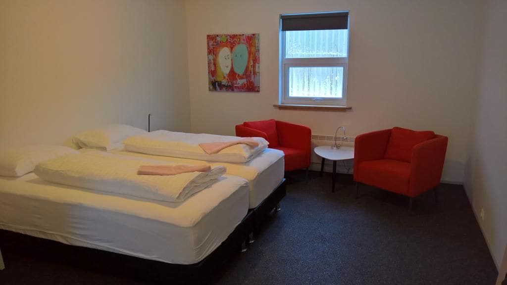 Ein Hotelzimmer mit zwei Betten, einem kleinen Tisch, zwei roten Stühlen, einem Fenster und einem farbenfrohen Gemälde an der Wand.