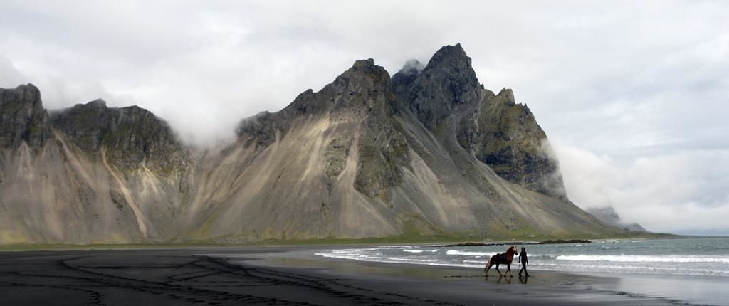 En strand med en hest og rytter, et bjerg i baggrunden og en overskyet himmel.