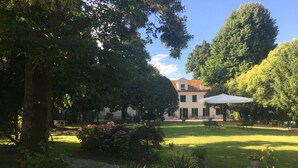 Un jardin bien entretenu avec un grand arbre, un bâtiment doté d’un toit recouvert de tuiles rouges et un parasol blanc.