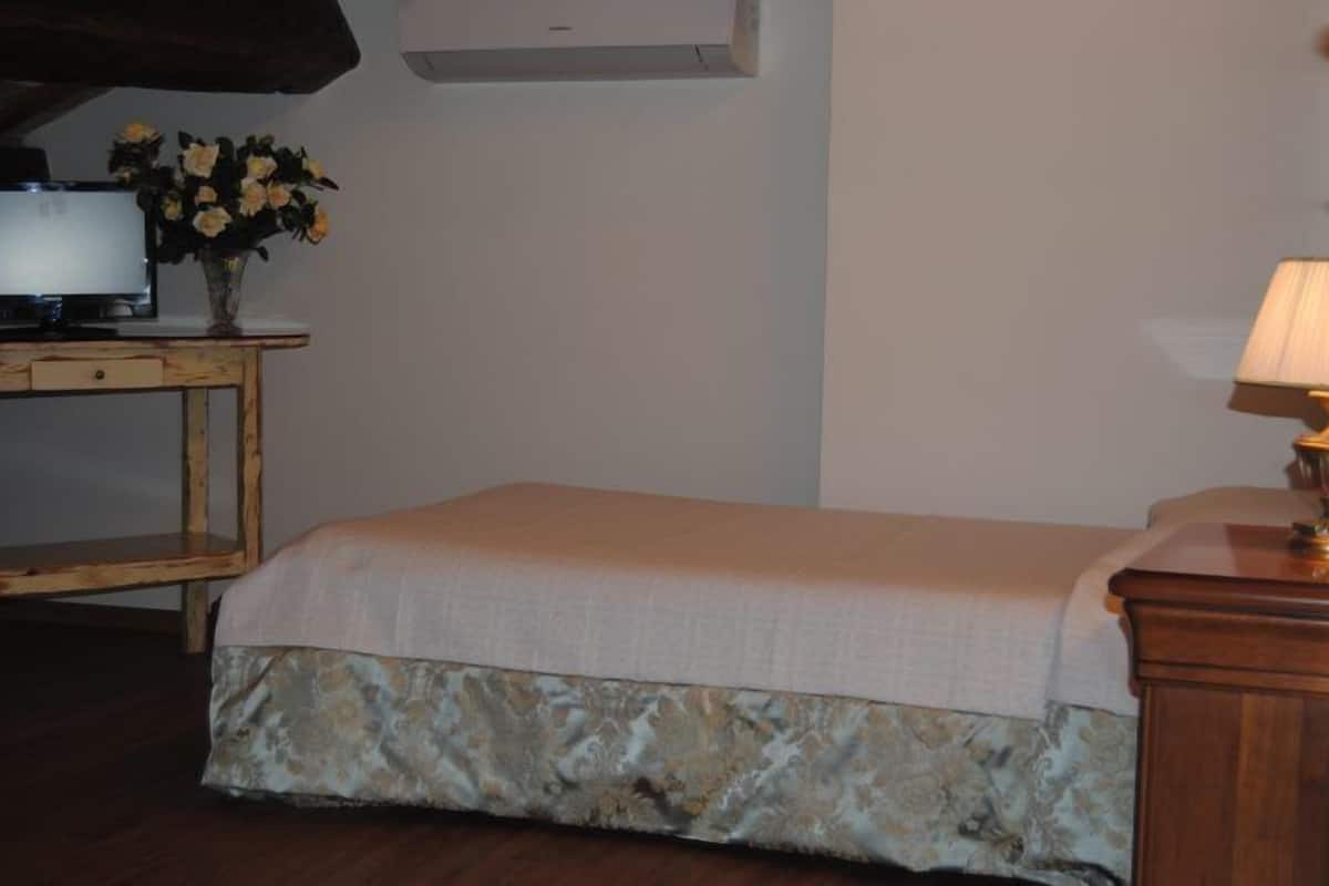 A bedroom with a bed, a wooden nightstand with a lamp, a TV, and a vase with flowers.