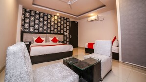 Une chambre d’hôtel moderne avec un grand lit, deux fauteuils blancs, une table basse en verre, un ventilateur de plafond et un papier peint à motifs.