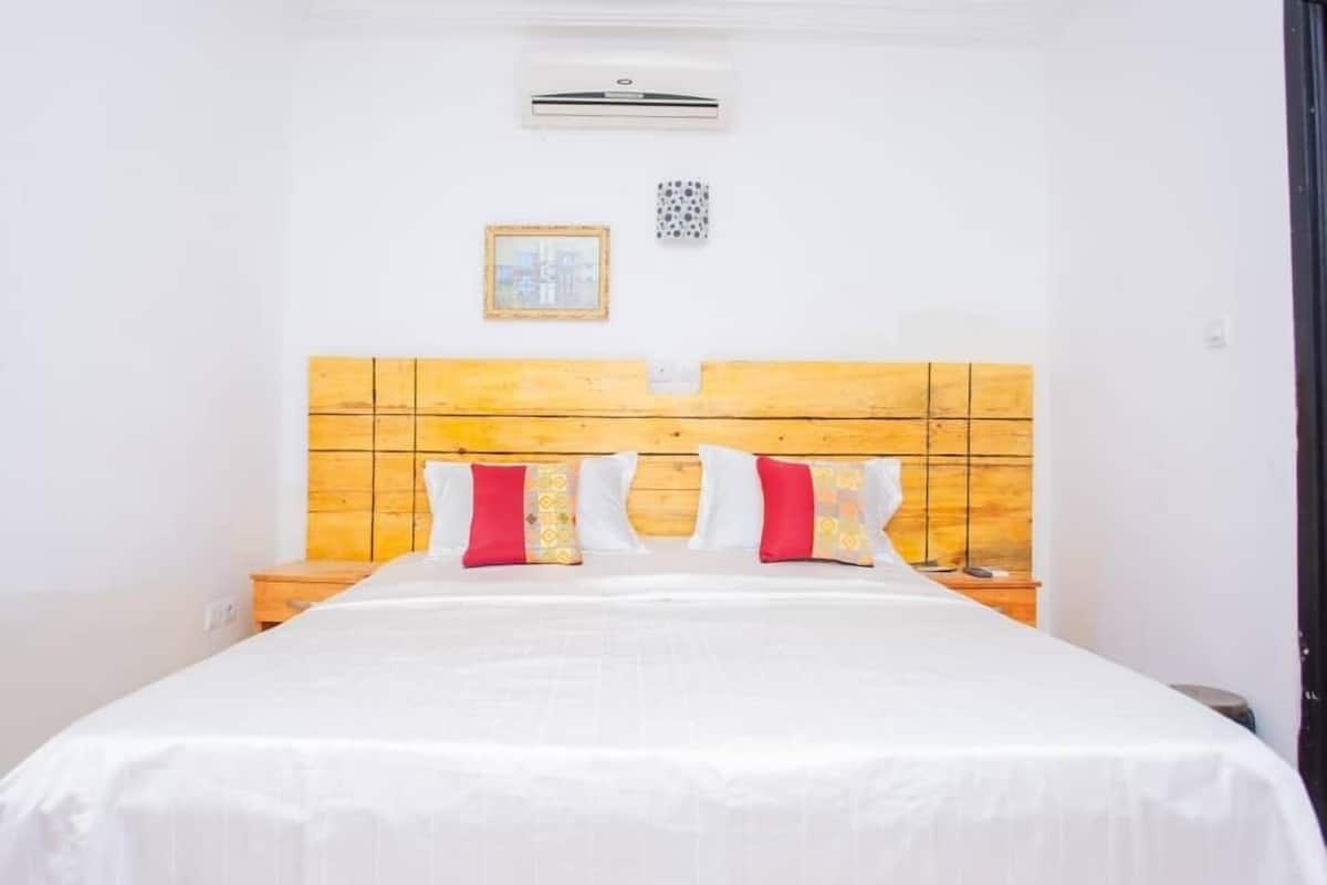A hotel room with a wooden headboard, a bed with white bedding and red and white pillows, a small bedside table, and a wall-mounted air conditioning unit.
