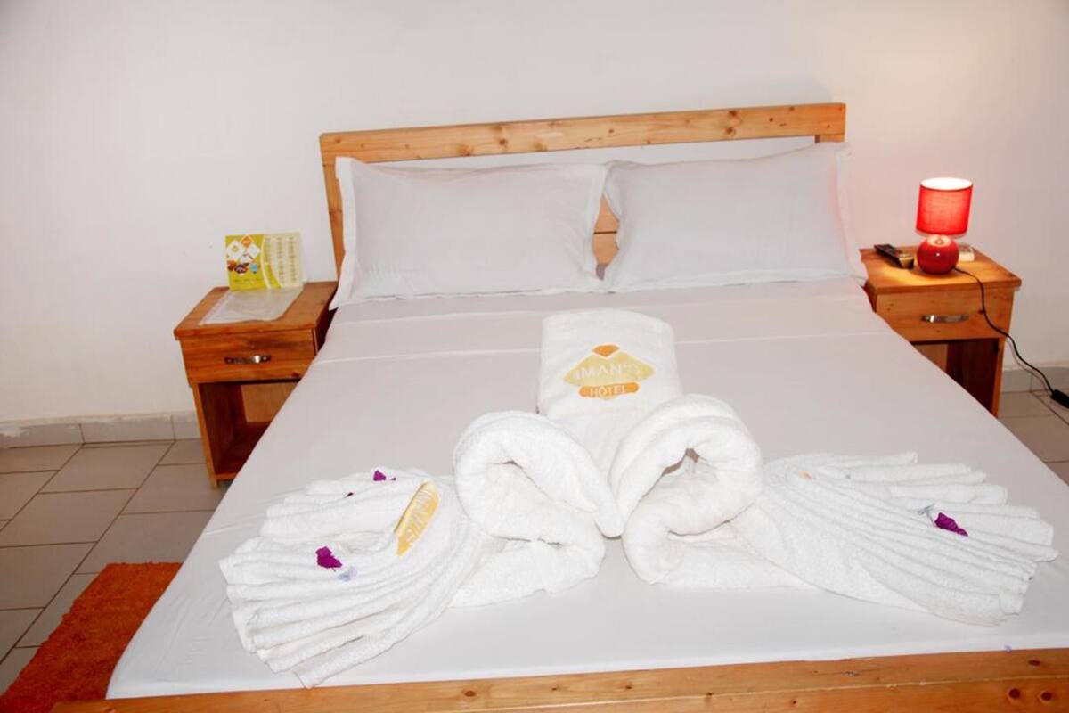 A neatly made bed with towels and a hotel logo, wooden headboard, white pillows, and a red lamp on a wooden nightstand.